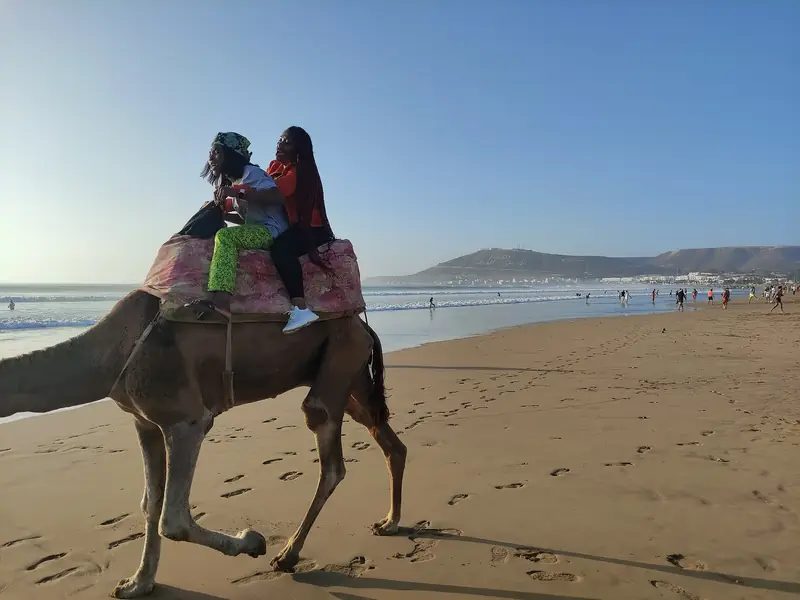Two visitors riding a camel along Agadir beach with the bay behind them