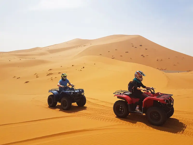 Two riders in helmets on quad bikes crossing golden sand dunes in Morocco