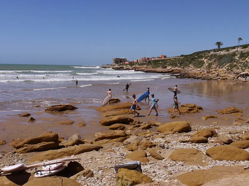 Surfers carrying boards across the rocky shore of Panorama beach in Taghazout